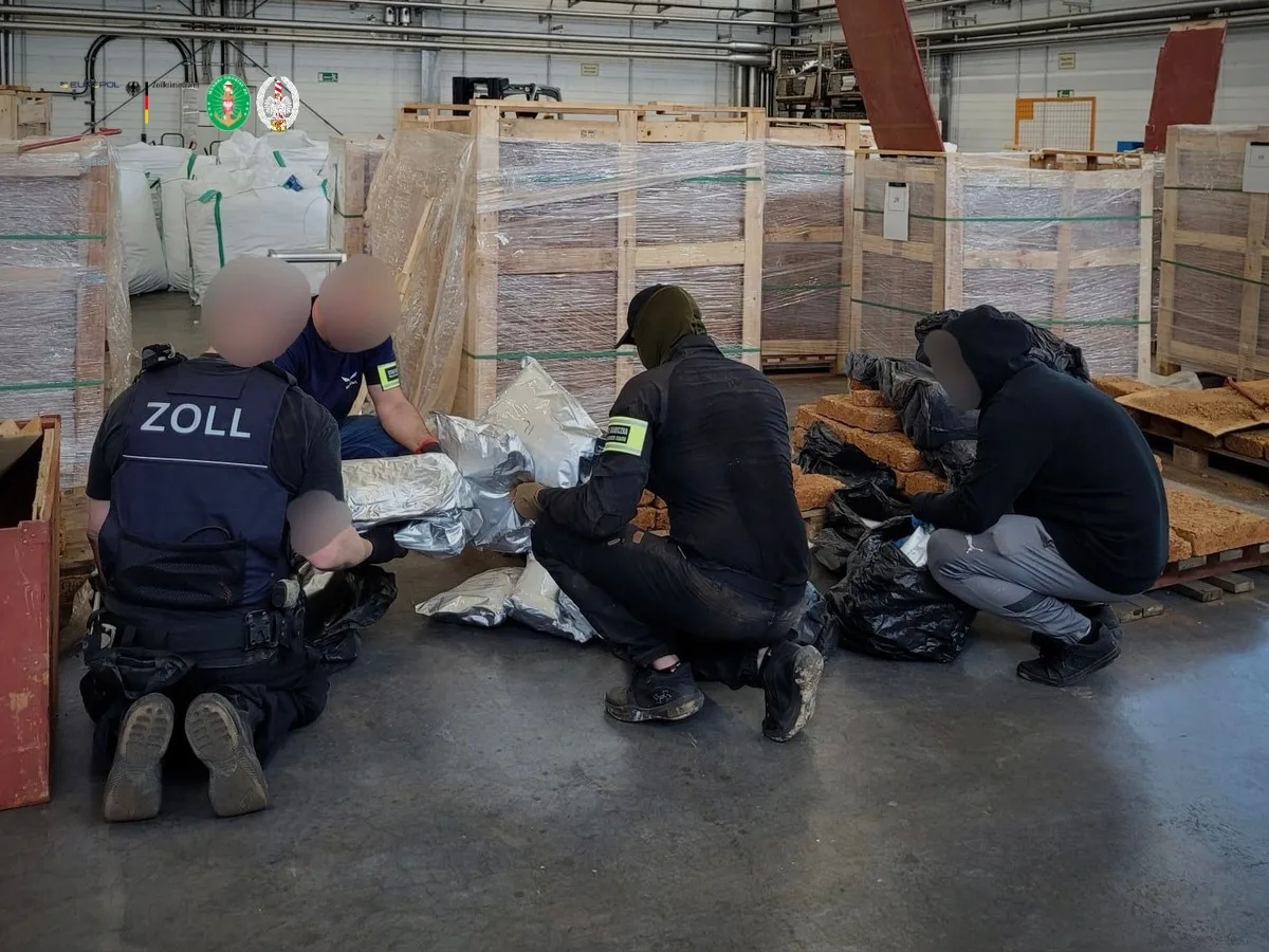 Vier Personen, darunter Zollbeamte, durchsuchen in einer Lagerhalle Kisten und säcke mit verdächtigen Inhalten. Kisten sind teilweise geöffnet, und die Beamten tragen Westen mit Aufschrift ‚Zoll‘.