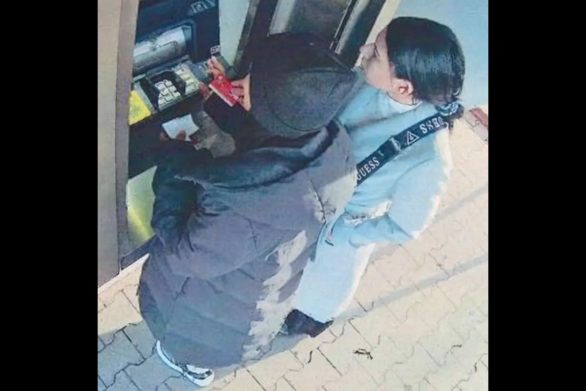 Zwei Personen stehen an einem Geldautomaten; die vordere Person trägt eine schwarze Jacke und bedient den Automaten.
