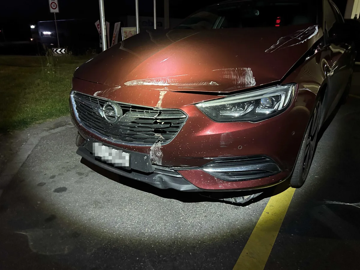 Ein rotes Auto mit sichtbarem Frontschaden steht nachts auf einer Straße bei schwacher Beleuchtung. Der Schaden ist besonders am Kühlergrill deutlich erkennbar.