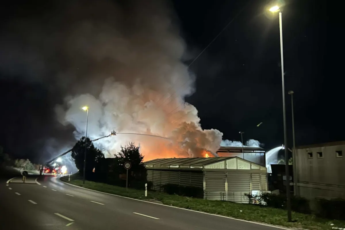 In der Nacht bildet sich über einem brennenden Industriegebäude dichter Rauch. Flammen sind hinter dem Gebäude zu erkennen.
