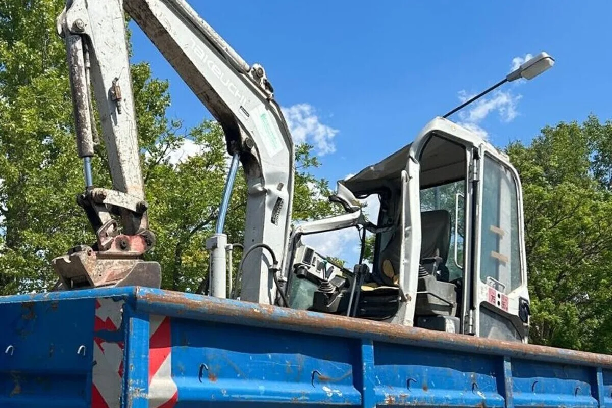 Ein kleiner, weißer Bagger ist auf der Ladefläche eines blauen Lastwagens abgestellt. Der Hintergrund zeigt Bäume und einen wolkenlosen blauen Himmel.