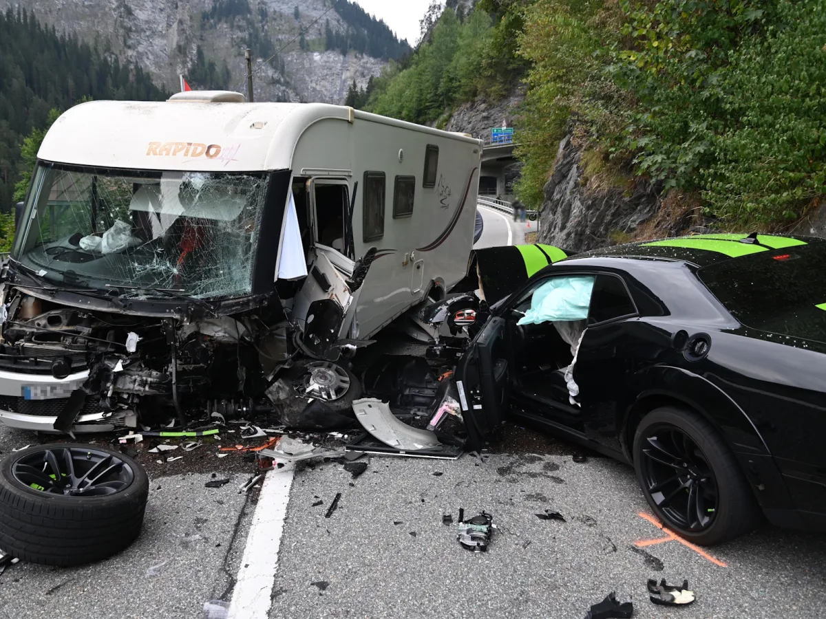 Ein Wohnmobil mit zerstörter Front und ein schwarzes Auto mit Airbag, beide zeigen erhebliche Schäden nach einer Kollision auf einer Bergstraße.