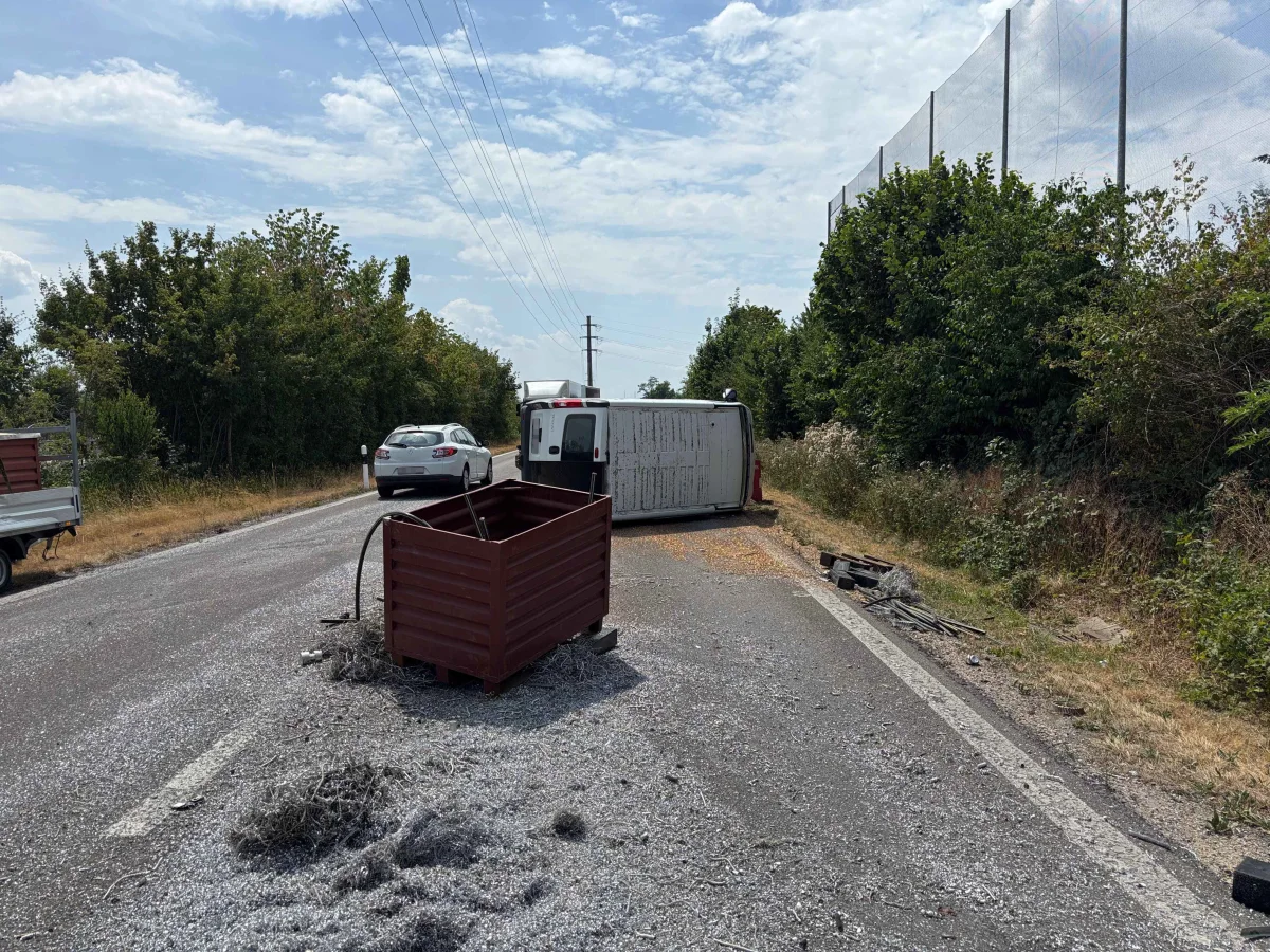 Auf einer Landstraße liegt ein umgekippter Anhänger, der Teile seiner Ladung verloren hat. Es sind auch andere Fahrzeuge und herumliegende Gegenstände zu sehen.