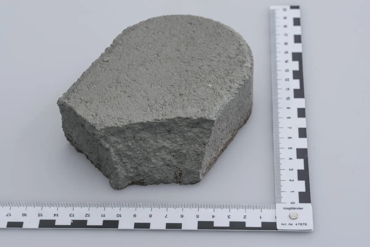 Ein graues, halbrundes Betonstück liegt auf einer glatten Oberfläche, flankiert von einem Maßband mit schwarzer und weißer Markierung.