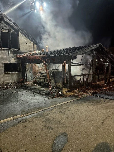 Ein stark beschädigter Carport mit einem ausgebrannten Fahrzeug steht neben einem stark zerstörten Gebäude. Rauch steigt auf.
