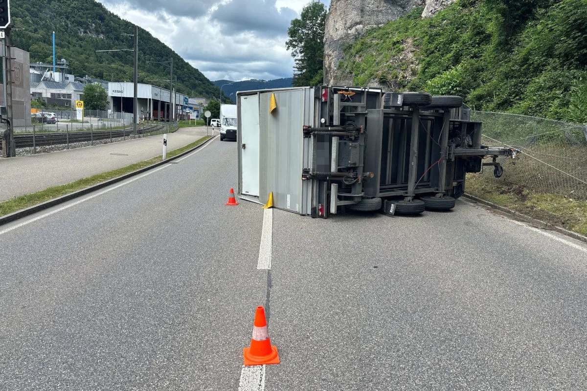 Ein umgekippter Anhänger liegt auf einer zweispurigen Straße, gesichert durch orange Absperrkegeln. Im Hintergrund sind Industriegebäude sichtbar.