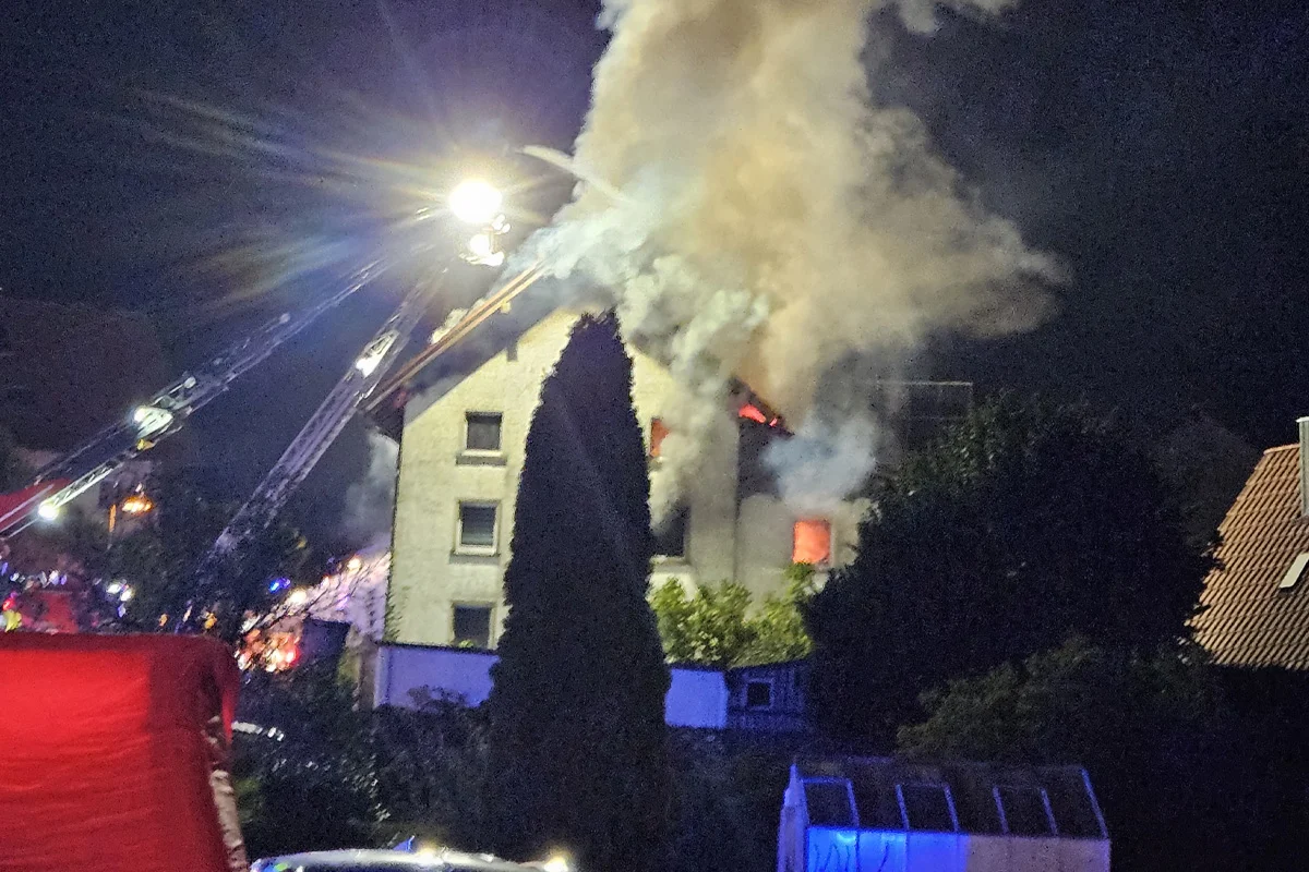 Ein Wohnhaus steht nachts in Flammen, während Feuerwehrkräne im Einsatz sind. Dicker Rauch steigt vom Dach auf.