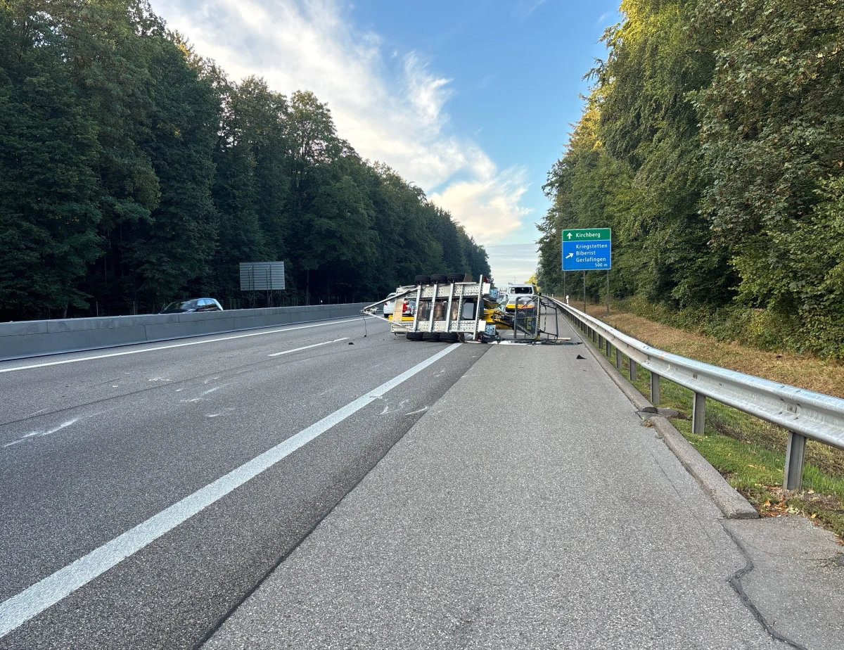 Ein umgekippter Anhänger liegt quer auf einer Autobahn und blockiert die Fahrbahn. Im Hintergrund ist ein Wald und ein Wegweiser zu erkennen.