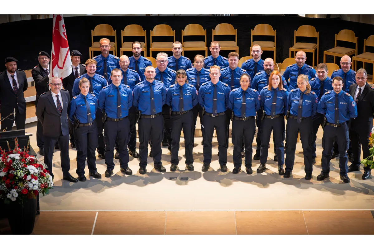 Eine Gruppe von Polizisten in blauen Uniformen steht in mehreren Reihen bei einer Zeremonie. Im Hintergrund sind leere Stühle zu sehen.