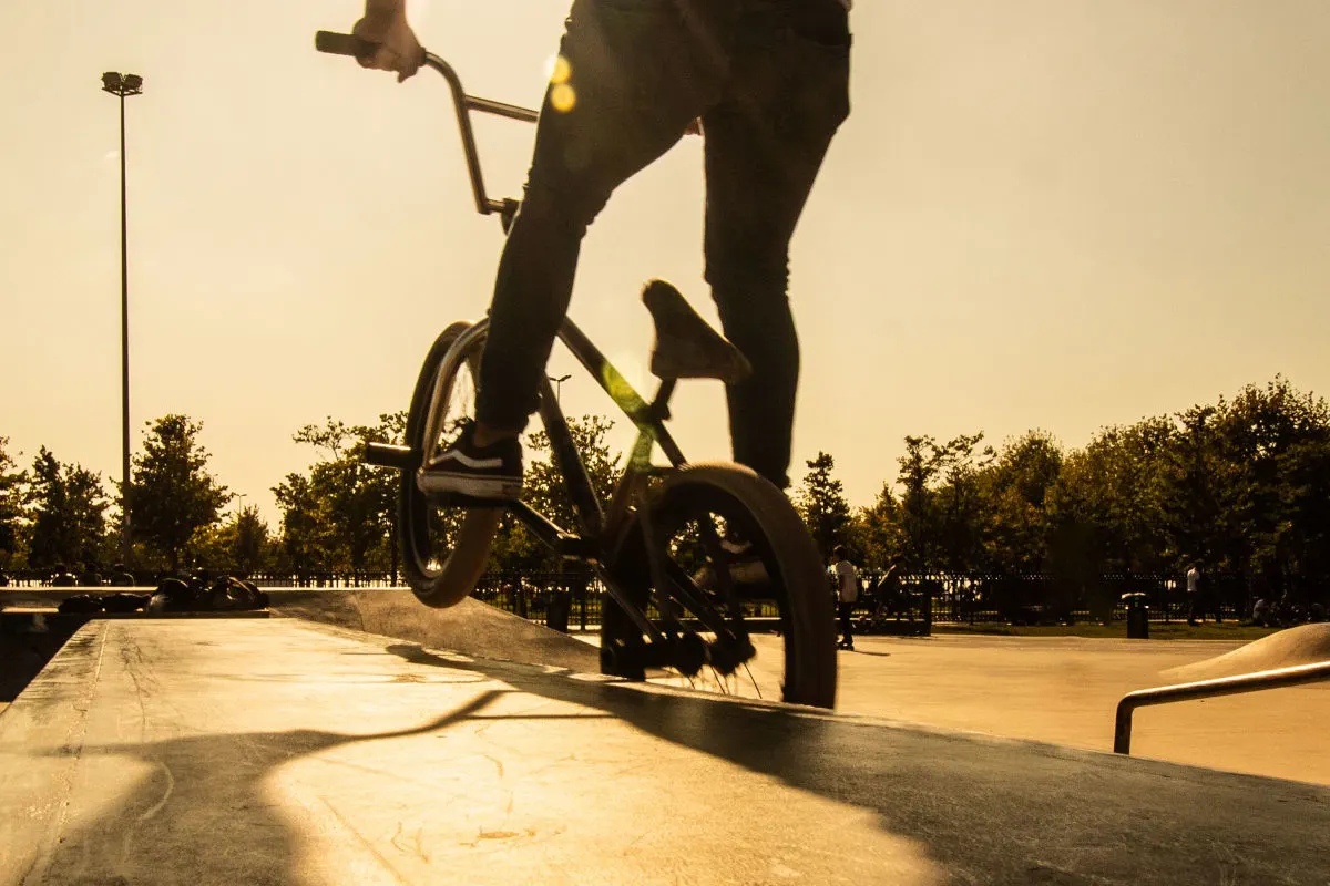 Ein BMX-Fahrer vollführt einen Trick auf einer Rampe, während im Hintergrund Bäume und weitere Personen im Park zu sehen sind. Die abendliche Sonne wirft lange Schatten über die Szene.