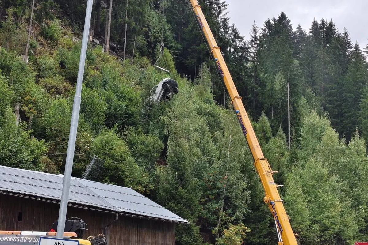 Ein weißes Auto hängt an einem Kran, der es aus einem dicht bewaldeten Hang zieht. Der Kran steht neben einer strukturierten Fläche.