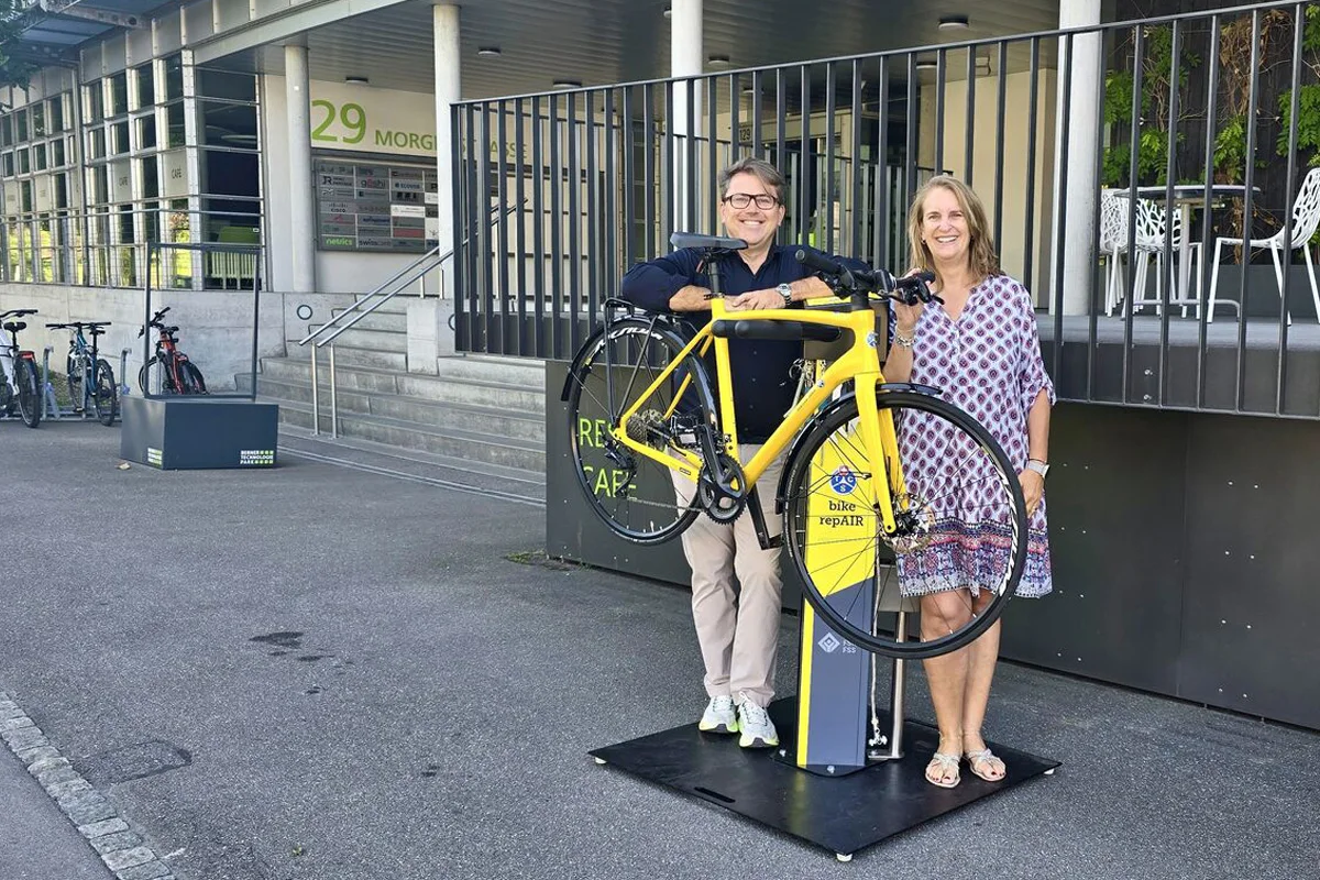 Eine Frau und ein Mann stehen neben einem gelben Fahrrad, das auf einer Reparaturstation befestigt ist, vor einem Gebäude.