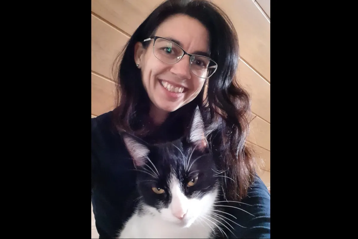 Eine Frau mit Brille und langen dunklen Haaren lächelt in die Kamera, während sie eine schwarz-weiße Katze hält.