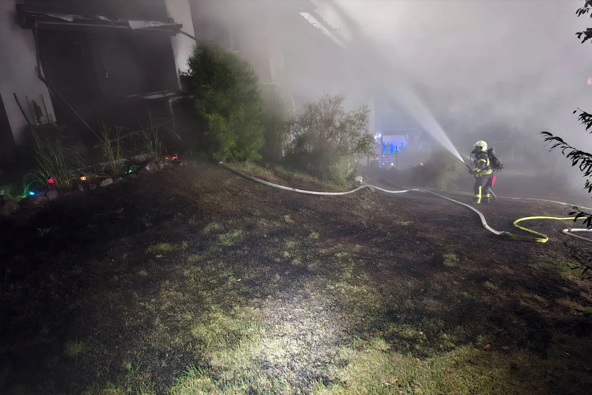 Ein Feuerwehrmann in Schutzkleidung bekämpft nachts mit einem Schlauch das Feuer an einem Wohnhaus. Dicker Rauch umgibt die Szenerie, das Umfeld ist unklar beleuchtet.