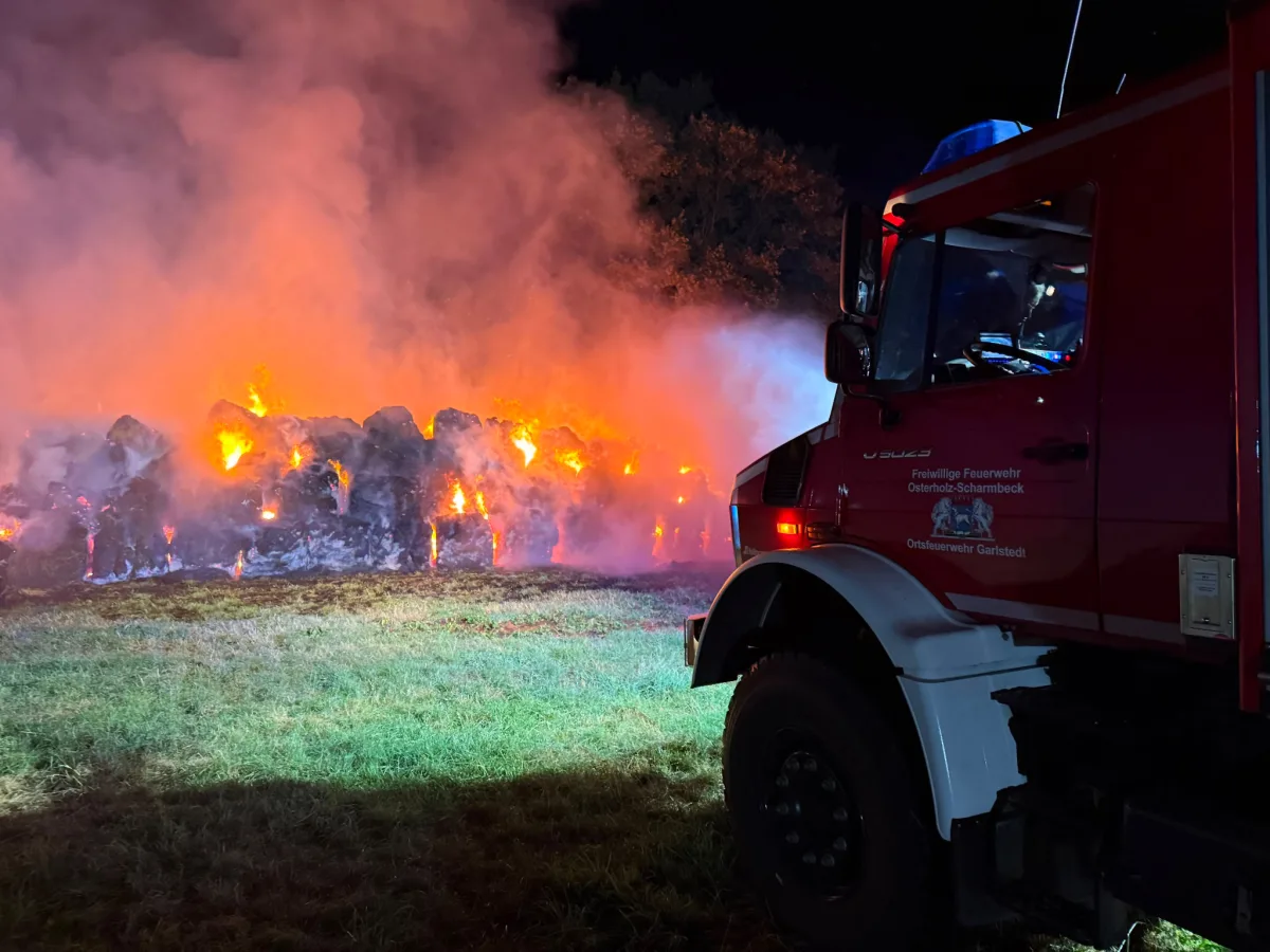 Ein rotes Feuerwehrauto der Freiwilligen Feuerwehr Osterholz-Scharmbeck steht neben einem großen Feuer aus brennenden Heuballen, das Nacht erleuchtet.