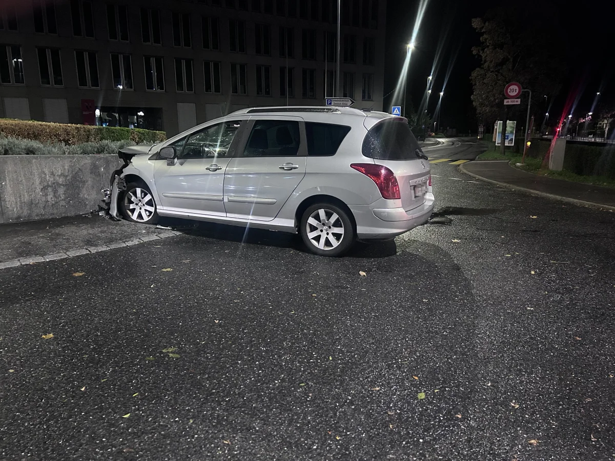 Ein silbernes Auto ist vorne stark beschädigt und steht gegen eine Betonmauer gelehnt. Die Szene ist nachts auf einer nassen Straße mit wenigen Beleuchtungen aufgenommen.