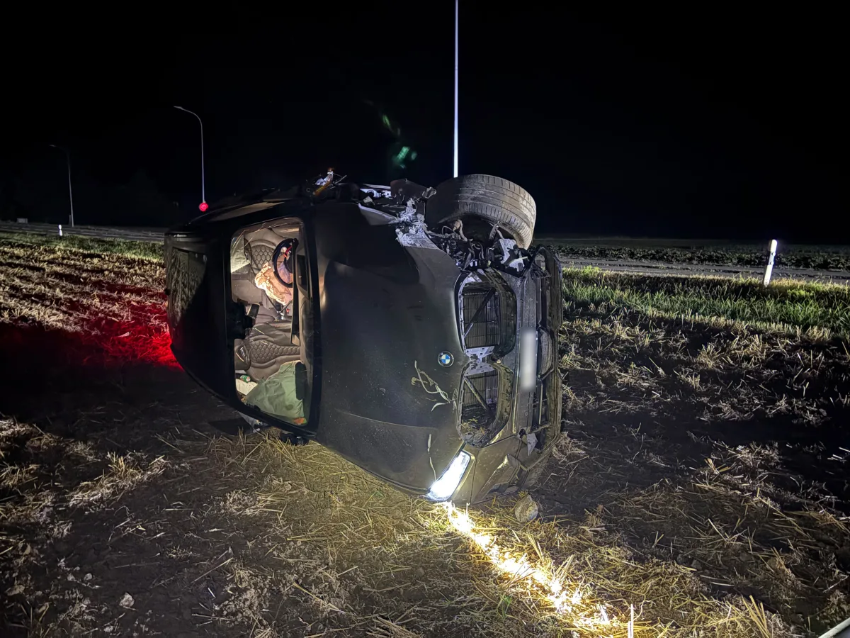 Ein schwarzer BMW liegt auf der Seite auf einem Feld bei Nacht. Das Auto ist beschädigt, mit geöffneter Beifahrertür, Scheinwerfer und der Unterboden sind sichtbar.