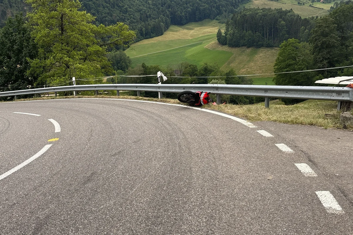 Ein rotes Motorrad liegt umgestürzt an einer Leitplanke in einer ländlichen Kurve, mit grünen Hügeln im Hintergrund.