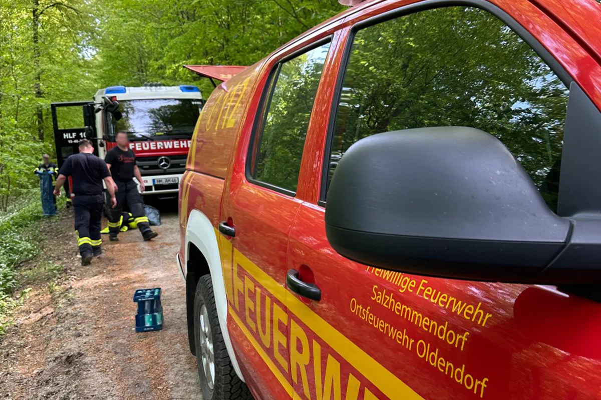 Zwei Feuerwehrfahrzeuge stehen auf einem Waldweg, umgeben von grünen Bäumen. Mehrere Feuerwehrleute in Uniform sind zu sehen.