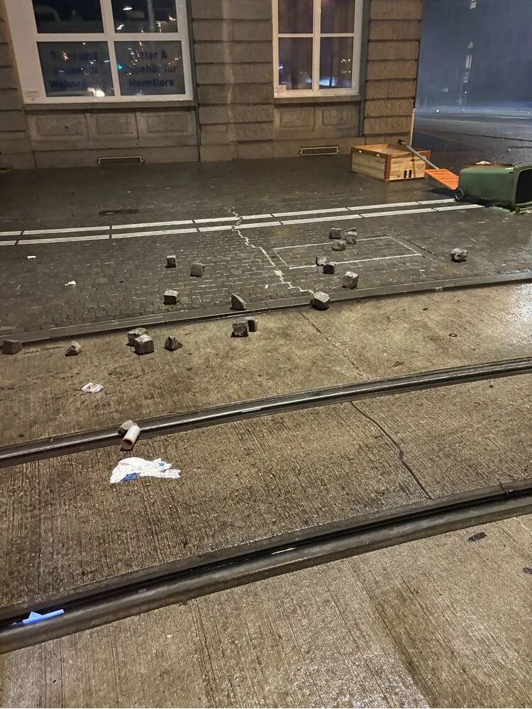 Auf einer gepflasterten Straßenszene liegen Pflastersteine verstreut, daneben ein umgestürzter Mülleimer. Die Szene spielt nachts.