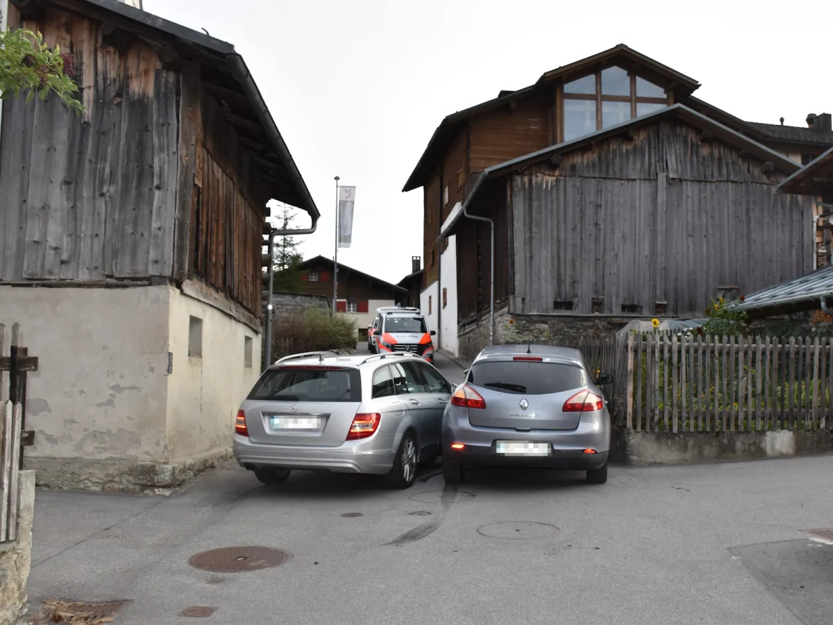 In einer engen Gasse zwischen Holzhäusern stehen zwei Autos dicht nebeneinander. Ein weiteres Fahrzeug steht dahinter.