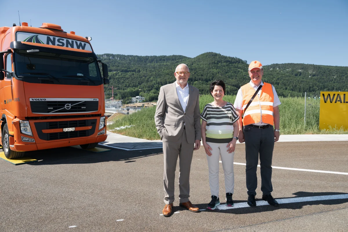 Drei Personen posieren vor einem orangefarbenen LKW auf einer neuen Straße. Im Hintergrund sind grüne Hügel erkennbar.