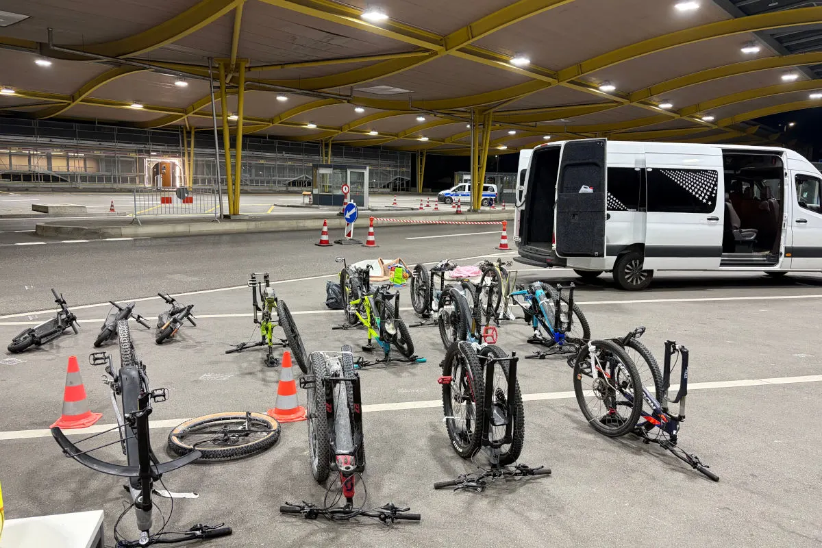 Mehrere Fahrräder liegen auf dem Boden vor einem geparkten Van mit geöffneten Türen. Der Schauplatz ist eine beleuchtete Kontrollstation in der Nacht.