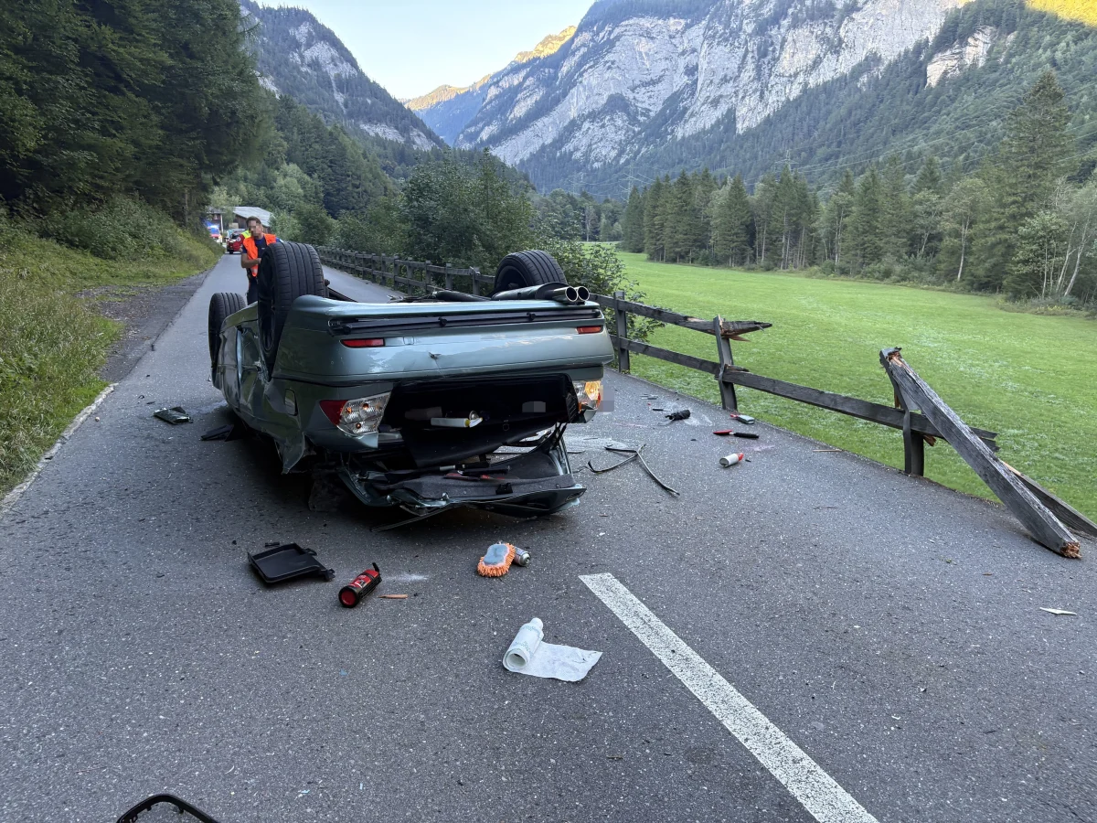 Ein Auto liegt auf dem Dach auf einer Straße, umgeben von verstreuten Gegenständen. Neben der Straße befindet sich ein beschädigter Holzzaun in einer bergigen Landschaft.