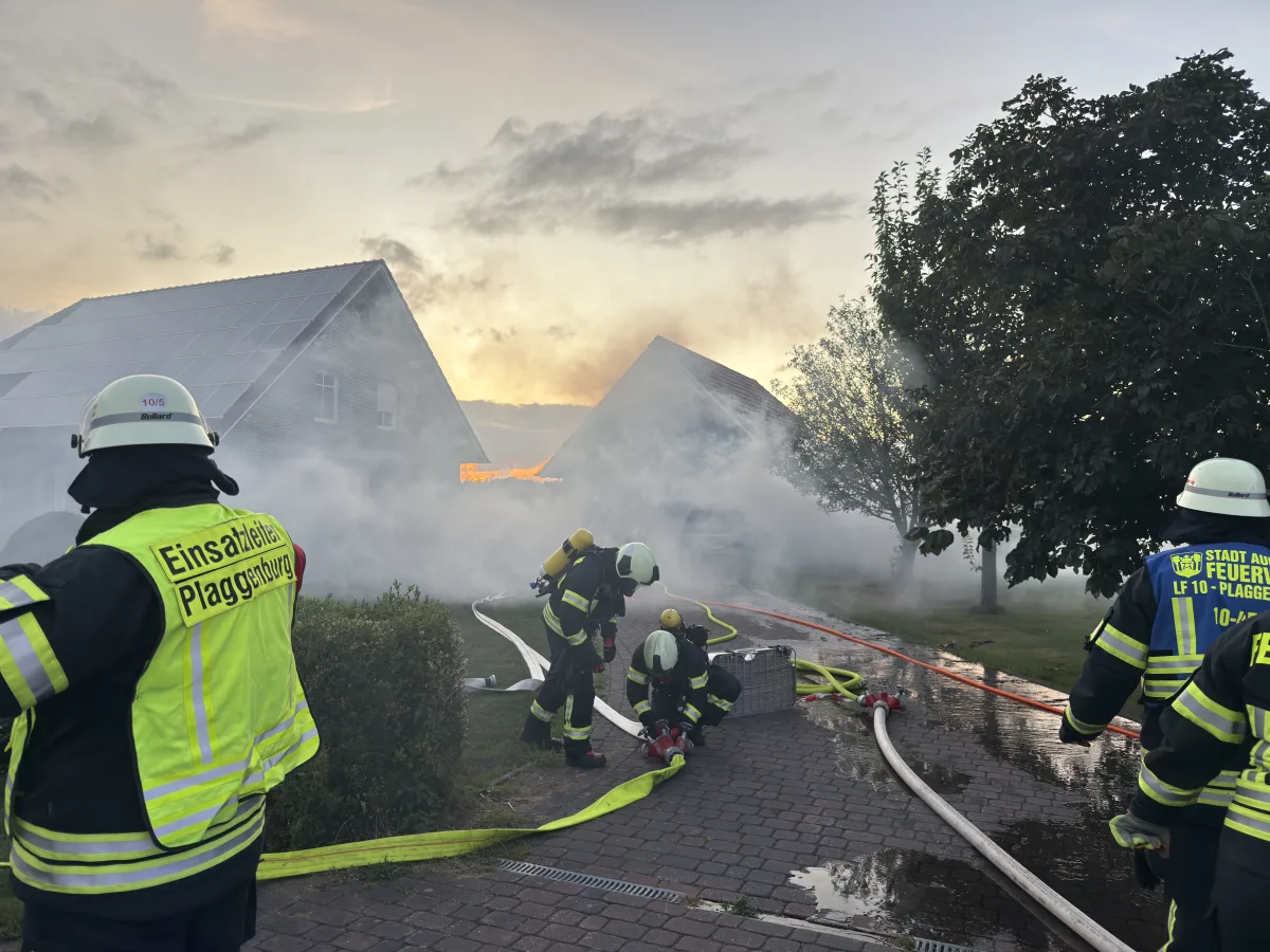 Mehrere Feuerwehrleute sind im Einsatz, um einen Hausbrand unter Kontrolle zu bringen. Rauch steigt aus dem Gebäude auf, während Schläuche auf dem Boden liegen.