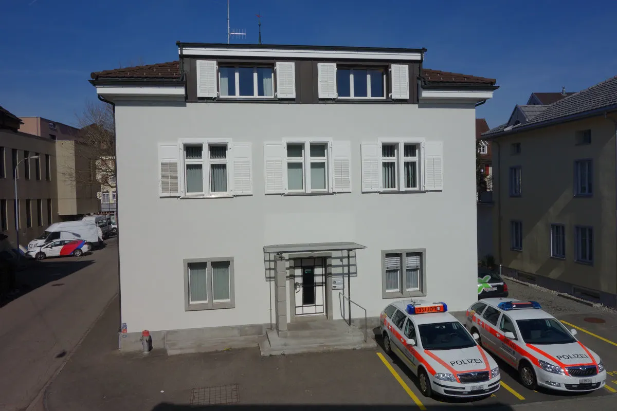 Das Bild zeigt ein weißes Gebäude mit zwei geparkten Polizeifahrzeugen davor. Im Hintergrund sind weitere Gebäude sichtbar.