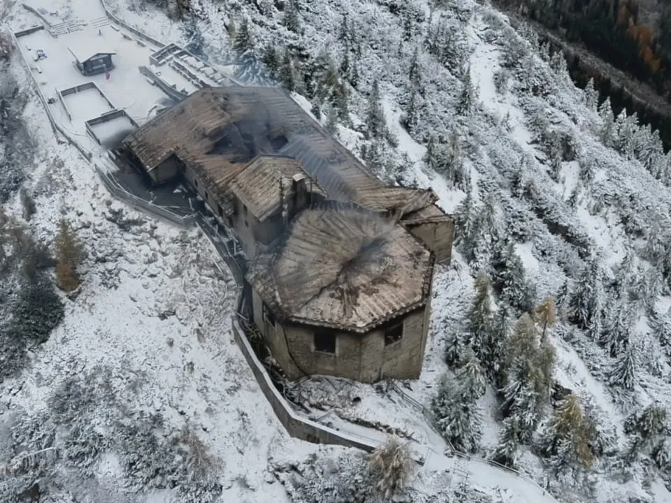 Ein großes, beschädigtes Gebäude befindet sich in einer verschneiten Berglandschaft, sichtbar aus der Vogelperspektive. Das Dach weist Spuren von einem Brand auf, umgeben von Bäumen und Schnee.