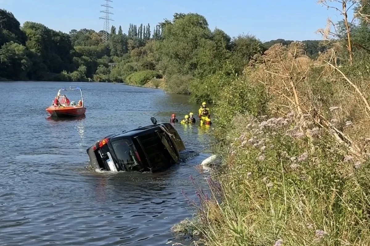Ein Auto liegt seitlich im Fluss, während mehrere Rettungskräfte in der Nähe im Wasser operieren. Im Hintergrund ist ein Rettungsboot zu sehen.