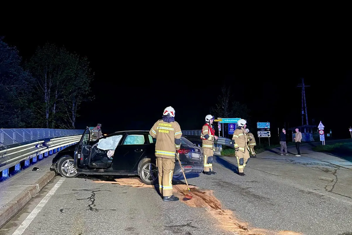 Ein schwarzes Auto mit beschädigter Vorderseite steht auf einer Straße. Feuerwehrleute in Uniform sind vor Ort und führen Aufräumarbeiten durch. Die Szene ist nachts beleuchtet.