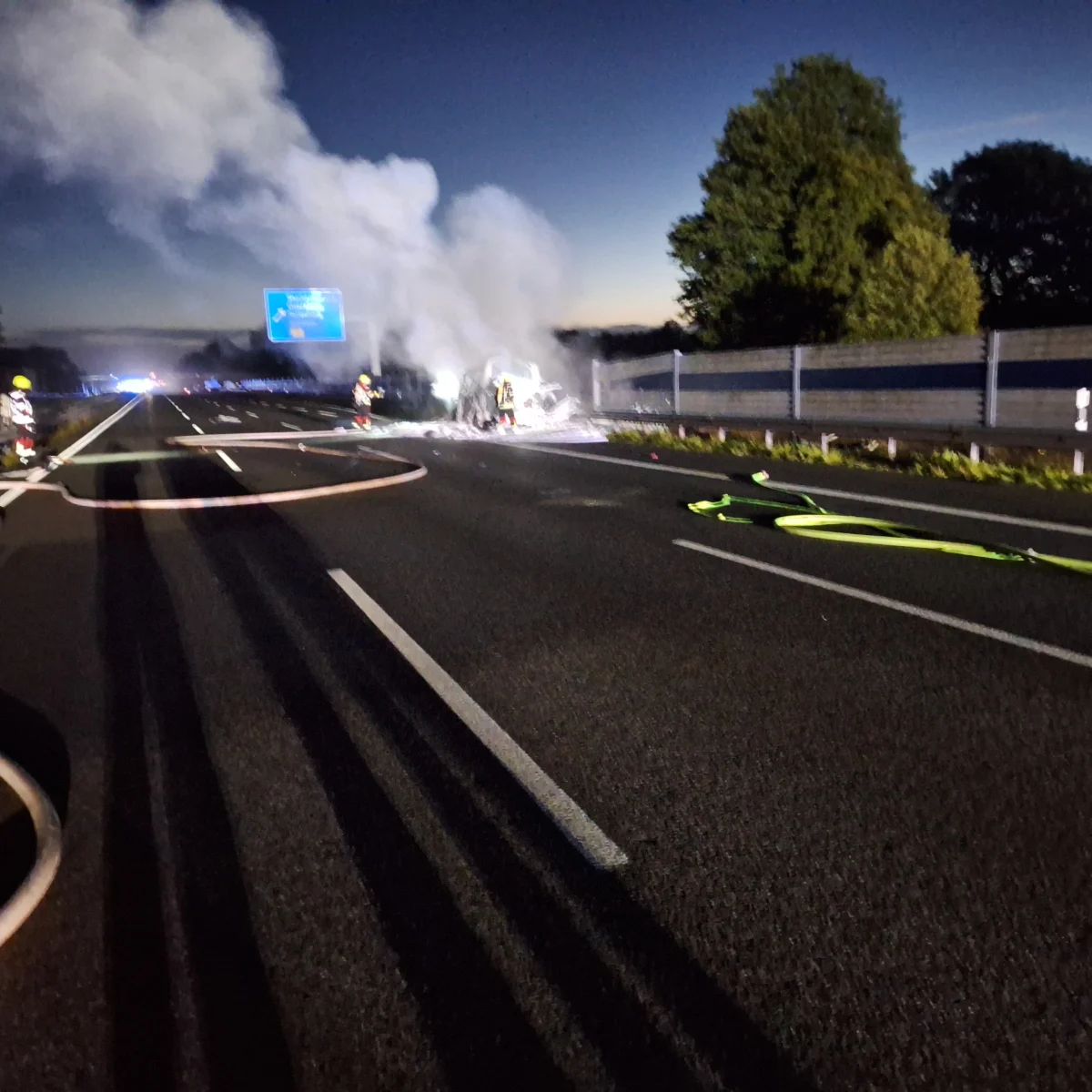 Mehrere Feuerwehrleute sind bei Nacht auf einer Autobahn zu sehen, während sie ein offensichtlich brennendes Fahrzeug löschen, das Rauch erzeugt.