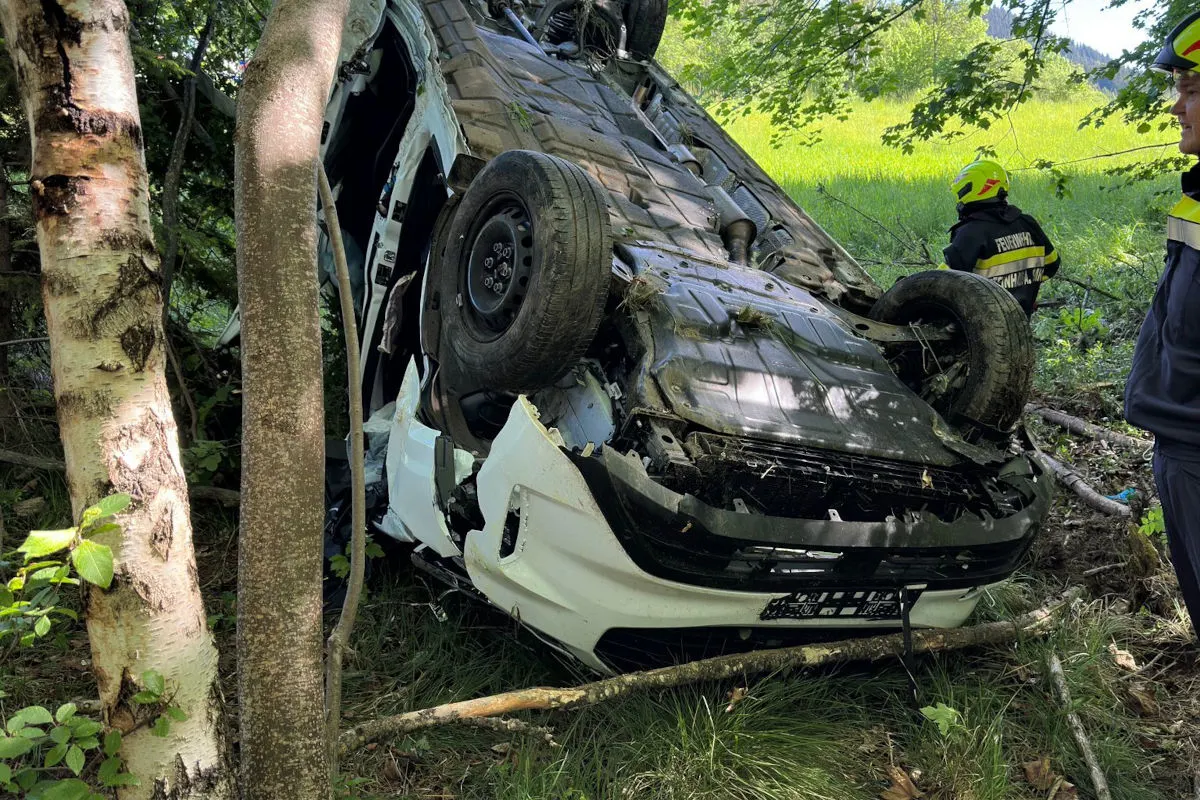 Ein weißes Auto liegt auf dem Dach unter Bäumen, umgeben von Gras und Ästen. Zwei Feuerwehrleute sind im Hintergrund sichtbar.