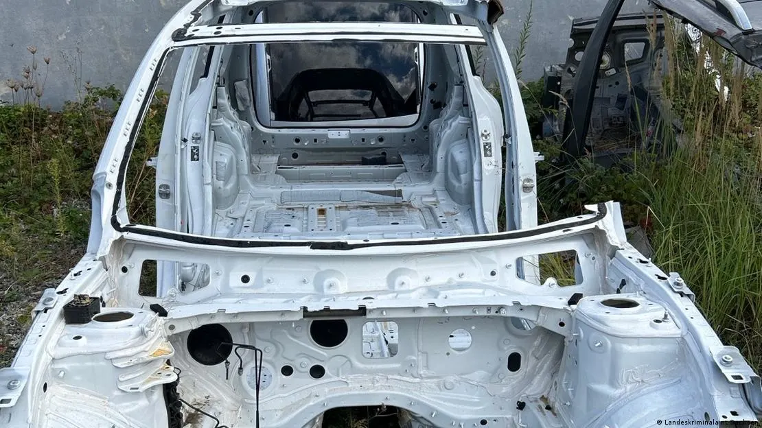 Ein ausgebranntes Autowrack ohne Türen und Fenster steht im Freien, umgeben von Gräsern und einer Betonwand.