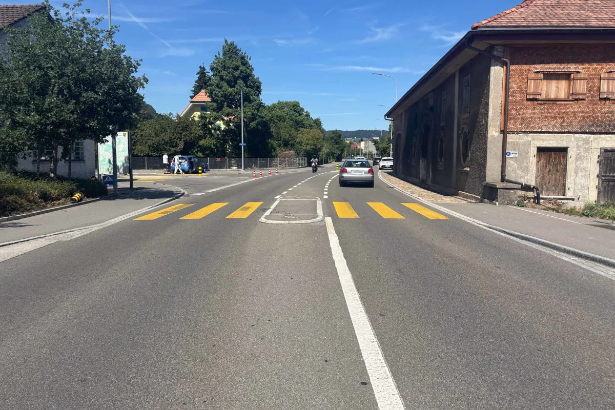 Eine Landstraße mit einem Zebrastreifen und mehreren parkenden Fahrzeugen ist bei klarem Himmel zu sehen. Am Straßenrand stehen Bäume und Gebäude.