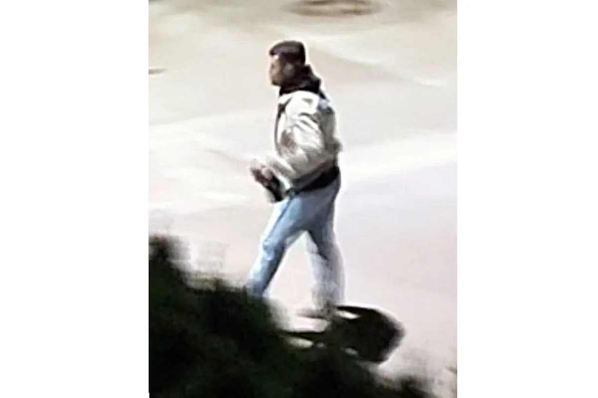 Ein Mann in einer hellen Jacke und blauen Jeans geht über eine Ansammlung von Asphalt bei Nacht. Ein Busch ist im Vordergrund zu sehen.