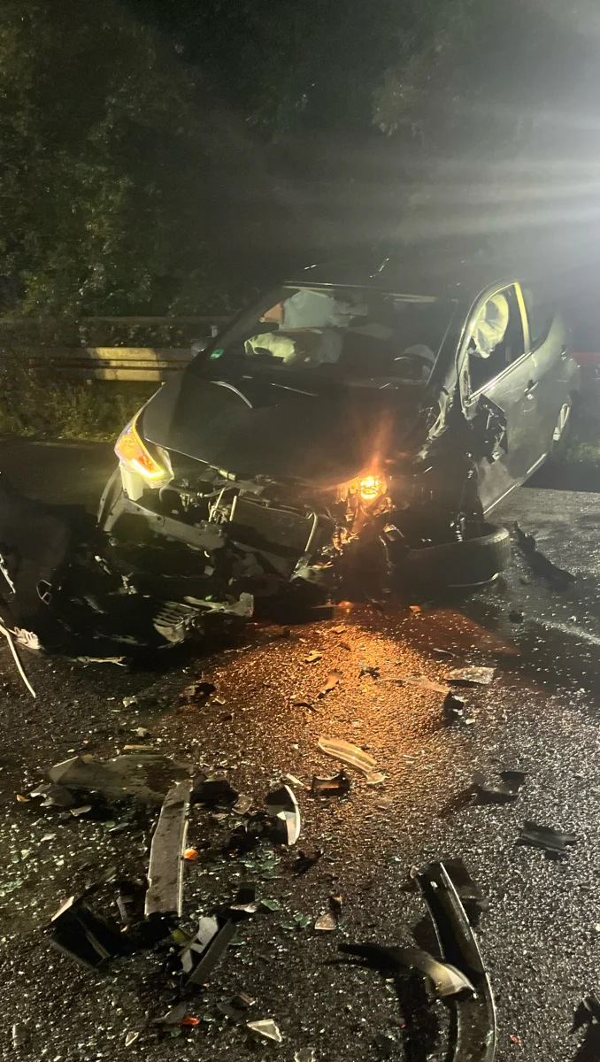 Ein schwarzes Auto mit stark beschädigter Front steht auf einer Straße, umgeben von Trümmern und Glasscherben. Die Szene ist bei Nacht aufgenommen und die Straße ist nass.
