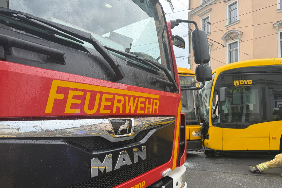 Ein rotes Feuerwehrfahrzeug ist im Vordergrund zu sehen, daneben steht ein gelber Bus mit einem 'DVB'-Logo. Es regnet.