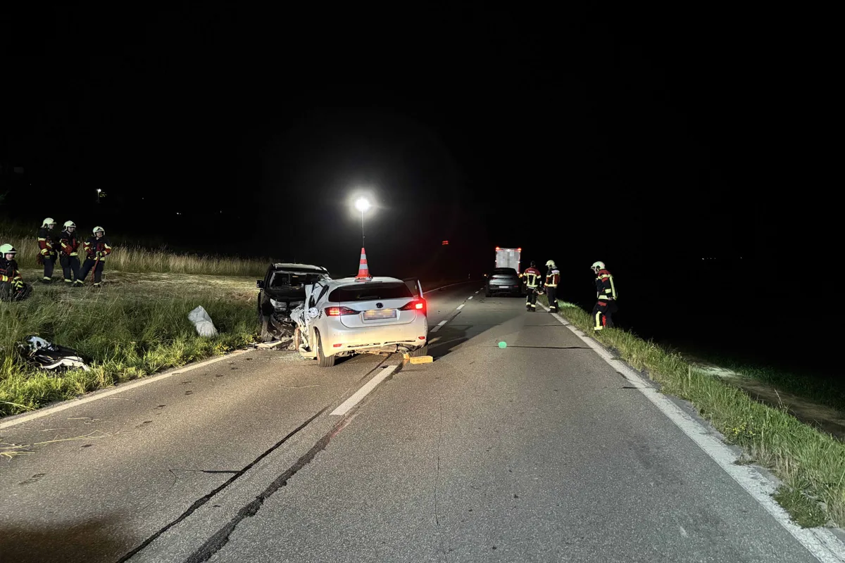 Eine nächtliche Szene zeigt ein beschädigtes Auto auf einer Landstraße, beleuchtet von einem Scheinwerfer. Feuerwehrleute sichern die Unfallstelle ab.