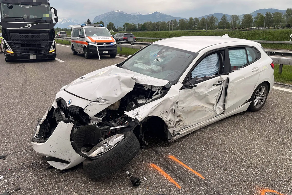Das Bild zeigt ein stark beschädigtes weißes Auto mit eingedrücktem Vorderteil auf der Autobahn. Im Hintergrund steht ein Polizeiwagen mit der Aufschrift 'Polizei' auf einem Streifenwagen.