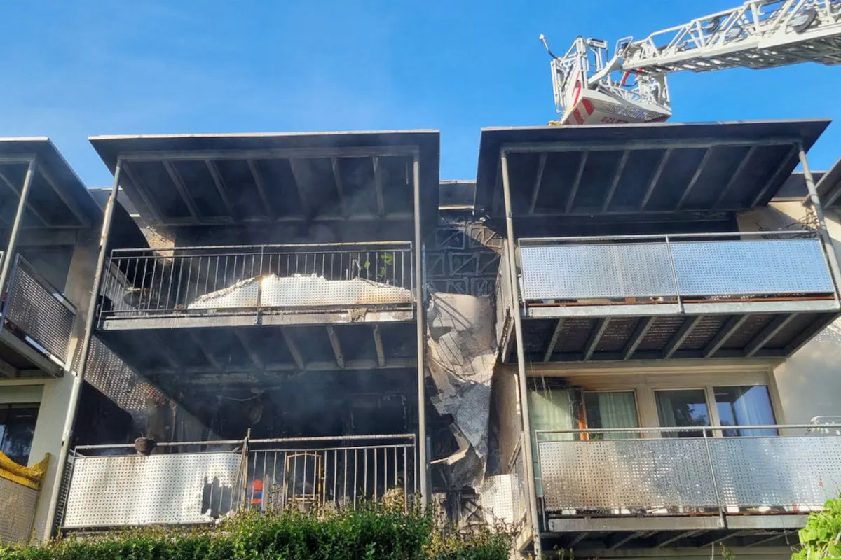 Ein Wohngebäude zeigt schwere Brandschäden an mehreren Balkonen, davon einer stark beschädigt. Eine Feuerwehrleiter ist im Einsatz.
