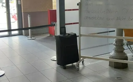 Ein schwarzer Koffer steht unbeaufsichtigt in einem öffentlichen Innenbereich neben einer Abgrenzung und einem Infoboard.