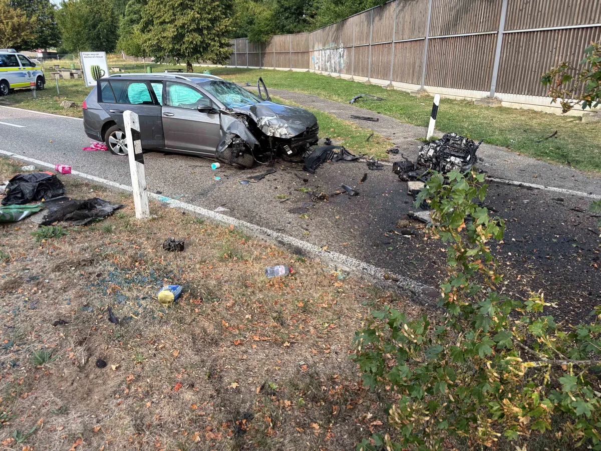 Ein graues Auto mit stark beschädigter Front steht schräg auf einer Landstraße. Trümmerteile sind auf der Straße verteilt.