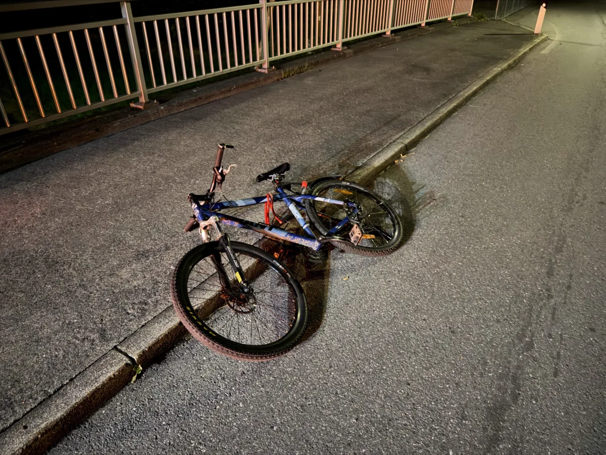 Ein Fahrrad liegt auf dem Boden einer beleuchteten Straße, neben einem Bürgersteig mit metallischem Geländer.