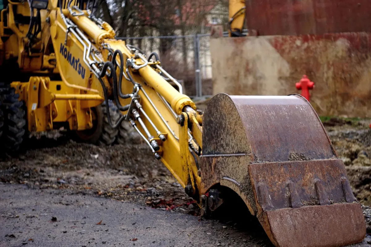 Ein gelber Baggerarm ragt in die Baustelle hinein, im Hintergrund ist ein roter Hydrant sichtbar. Der Boden ist erdig und uneben.