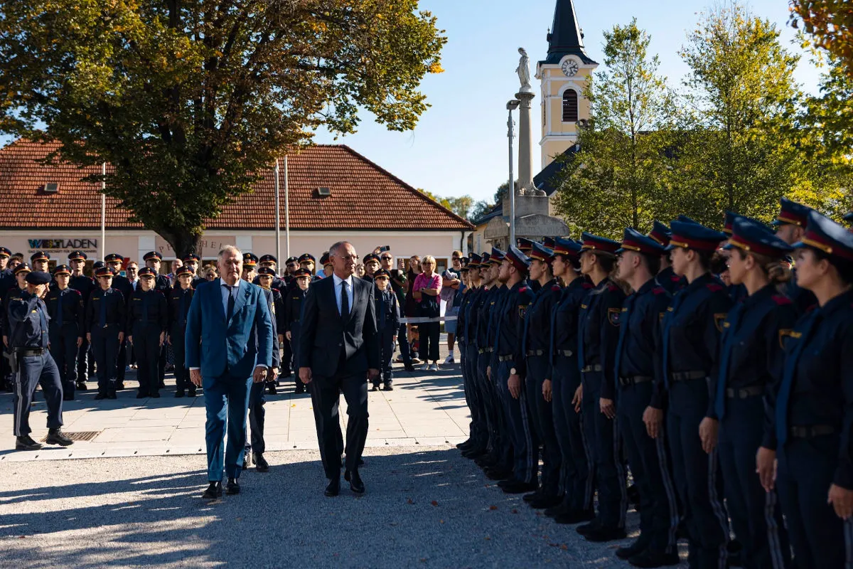 Zwei Herren in Anzügen führen eine Inspektion inmitten uniformierter Polizeischüler auf einem sonnigen Platz durch. Im Hintergrund ist eine Kirche zu sehen.