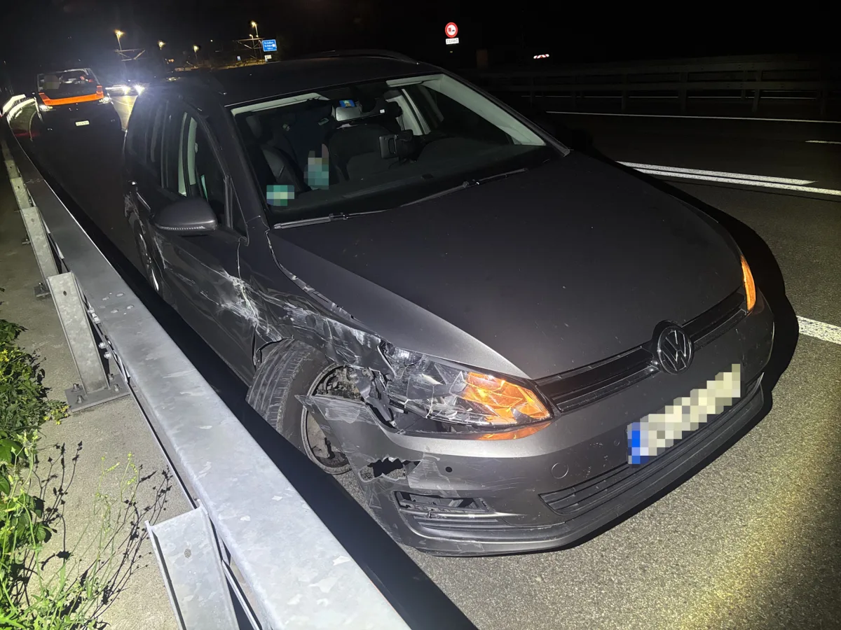 Ein dunkler VW mit Beschädigungen an der Front steht nachts auf einer Straße neben einer Leitplanke. Im Hintergrund sind weitere Fahrzeuge zu sehen.