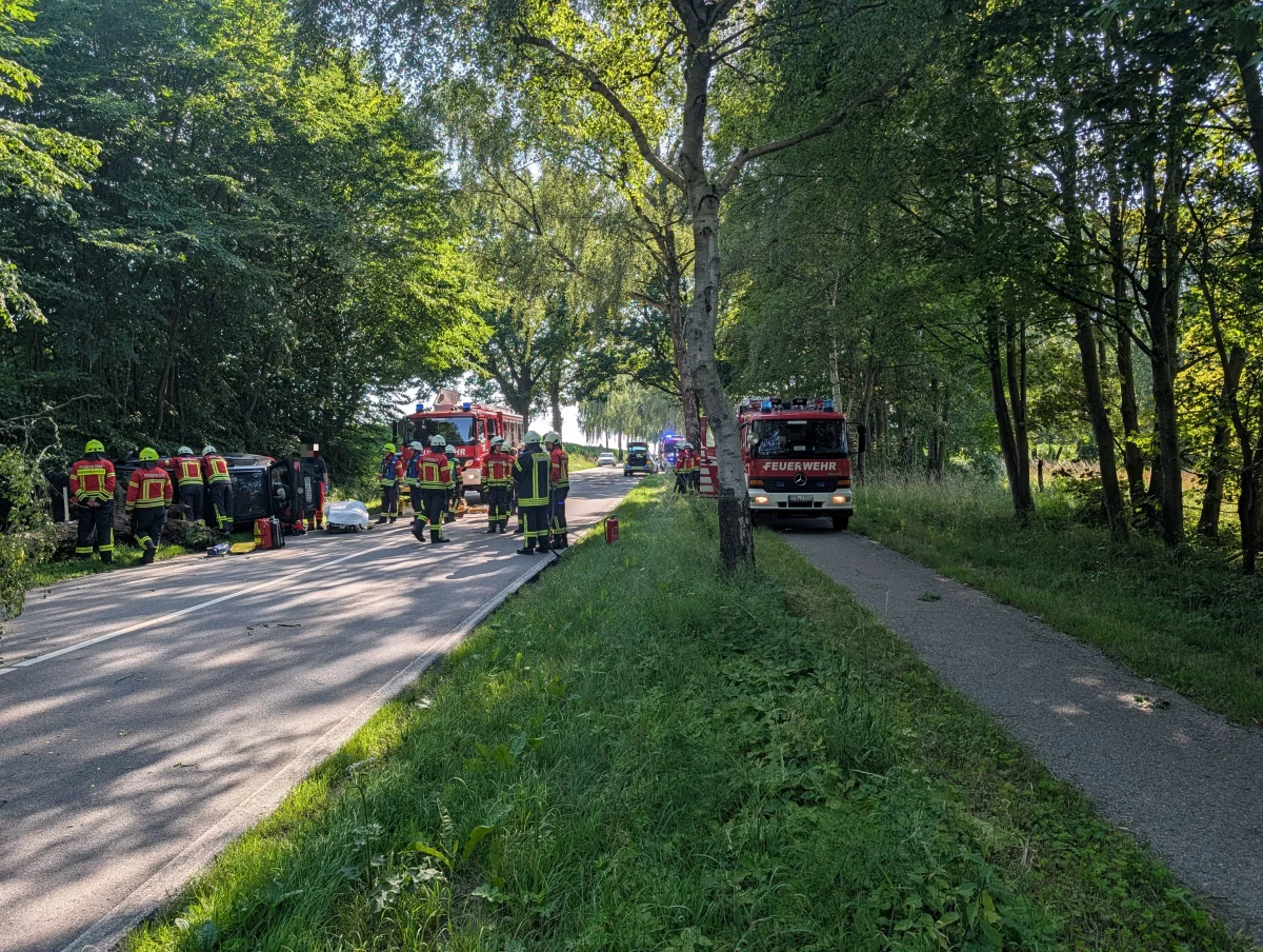 Ein umgekipptes Auto liegt seitlich auf einer Landstraße, umgeben von Feuerwehrleuten in Schutzkleidung. Im Hintergrund sind Feuerwehrfahrzeuge zu sehen.